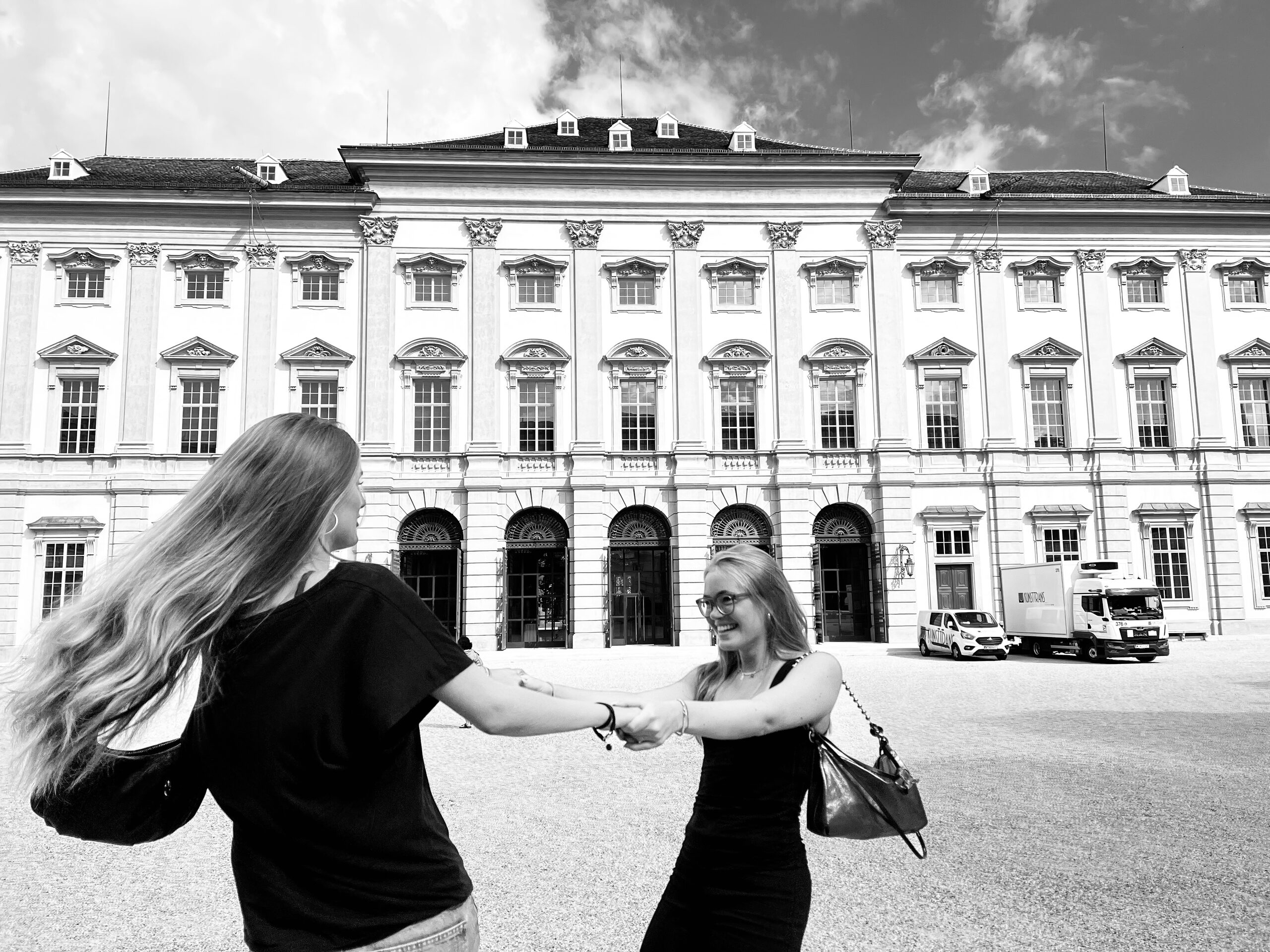 Zwei Frauen drehen sich Hand in Hand vor der langen barocken Fassade eines Palais.