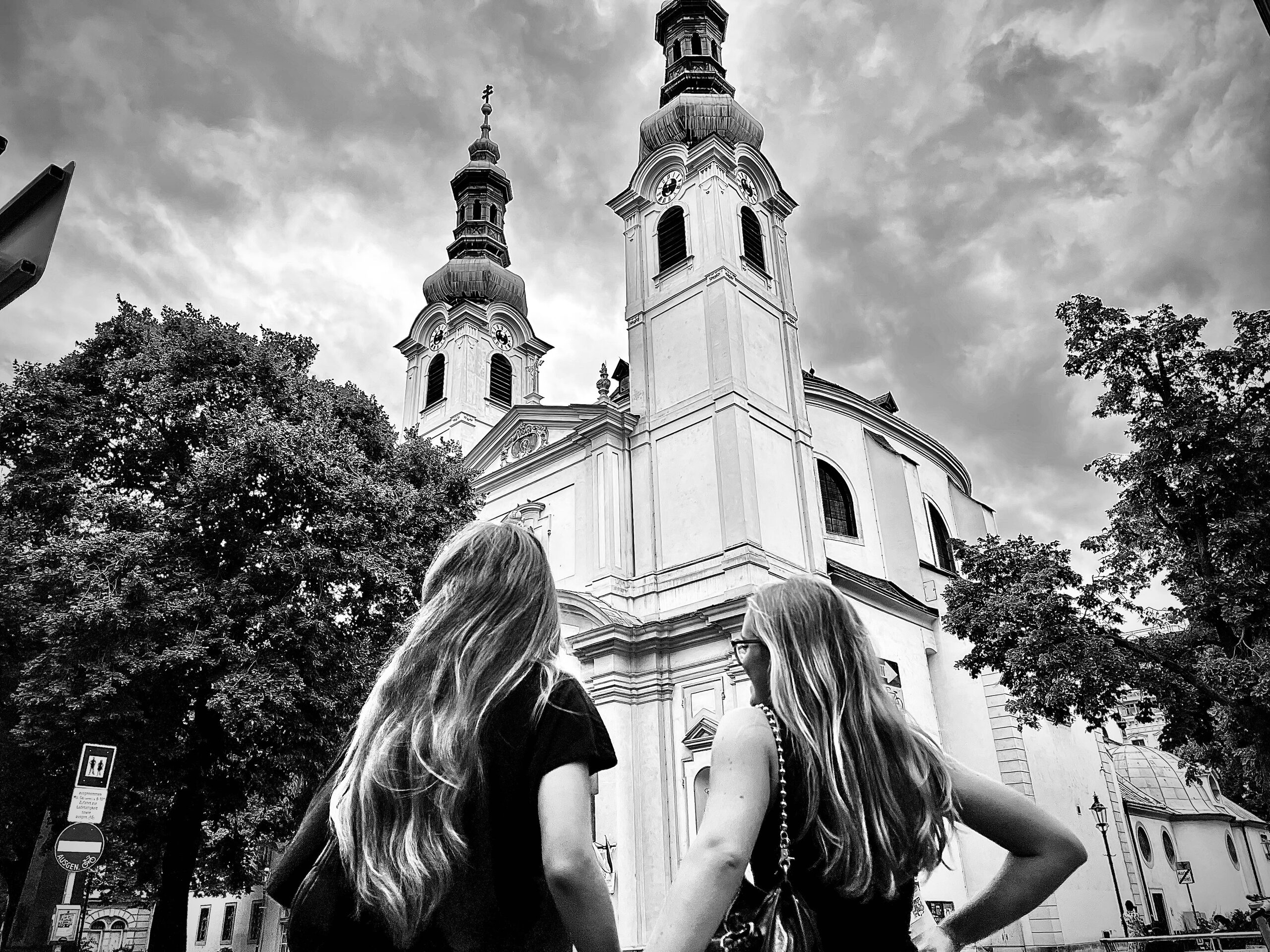 Eine barocke Kirche mit zwei Türmen, davor zwei Frauen unter den Bäumen.